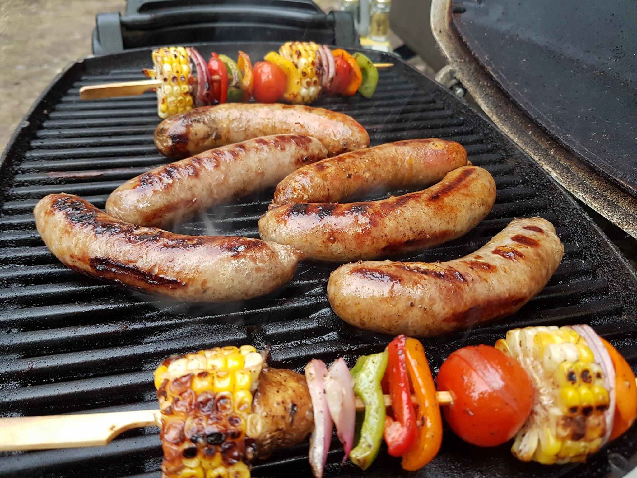 Photo of some sausages and vegetable skewers being cooked on a portable grill